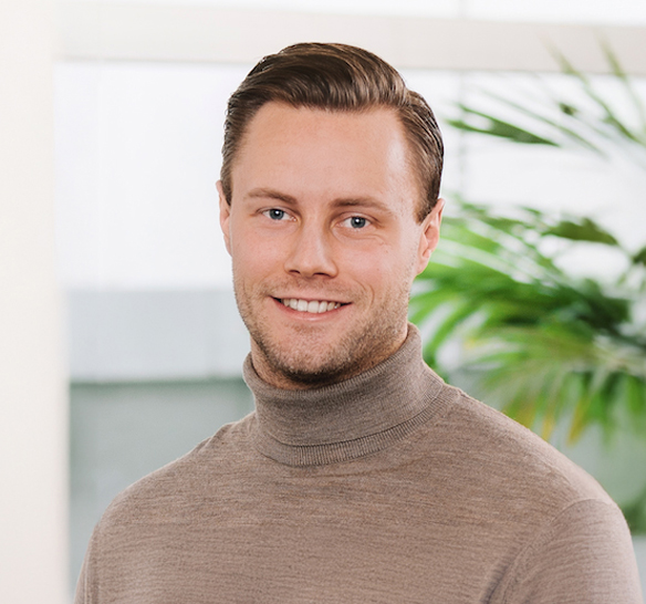 En leende man med kort, mörkt hår står framför en ljus bakgrund med gröna växter. Han bär en beige polotröja och tittar rakt mot kameran i en professionell porträttbild.
