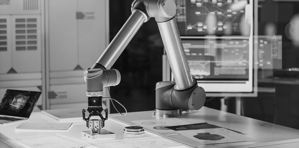 Robotarm med gripverktyg arbetar på ett bord fullt av tekniska ritningar och scheman. I bakgrunden syns skärmar med kod och data, vilket antyder en modern forsknings- eller utvecklingsmiljö. Arbetsytan är välordnad och ljus.