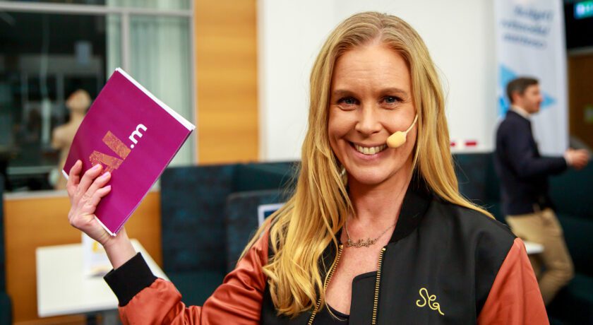 Anna Petersson. En kvinna med långt blont hår ler mot kameran. Hon har på sig en svart och orange bomberjacka med brodyr på vänster sida. Hon håller upp rosa talkort med texten AI.m på i höger hand. Hon har också en mikrofon fastsatt vid örat. I bakgrunden syns en suddig man i kavaj och ett modernt kontorsrum.