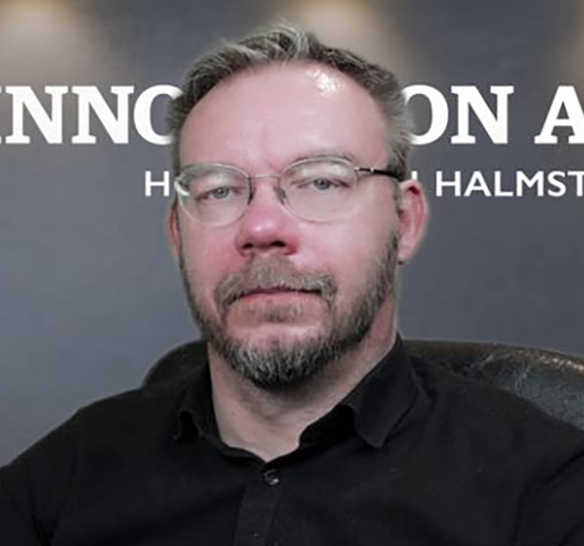 En man med kort hår, skägg och glasögon sitter framför en mörk bakgrund med vit text som delvis är synlig bakom honom. Han bär en svart skjorta och tittar rakt mot kameran med ett neutralt uttryck.