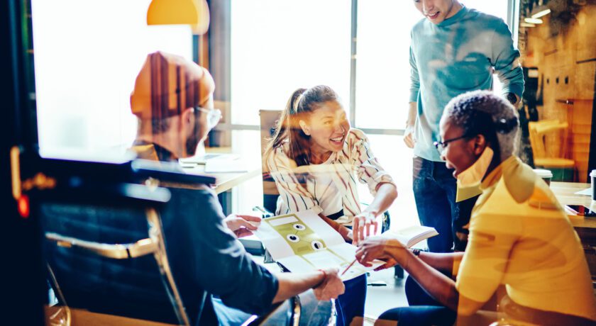 Fyra personer sitter och står runt ett bord i ett mötesrum med glasväggar. De tittar på dokument och skrattar tillsammans.
