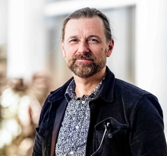 En man med skägg och långt hår i hästsvans står inomhus och tittar in i kameran. Han bär en mönstrad skjorta och en mörk jacka med ett par glasögon hängande i bröstfickan. Bakgrunden är ljus och suddig.