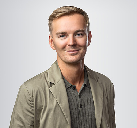 En man med kort blont hår ler lätt mot kameran. Han bär en ljusbeige kavaj över en mörkgrå ribbstickad tröja med knappar. Bakgrunden är ljusgrå och neutral.