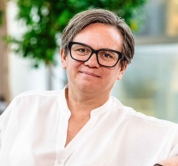 Porträtt av en kvinna med mörka glasögon. Hon bär en vit skjorta och ler. Bakgrunden är ljus med suddiga gröna växter, vilket ger ett fräscht och professionellt intryck.