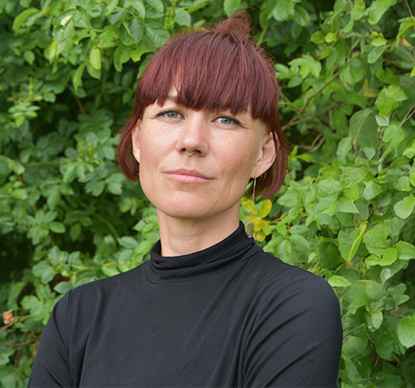 En kvinna med rödbrunt hår uppsatt i en knut och lugg står framför en grön, lövrik buske. Hon bär en svart, långärmad polotröja och har armarna i kors. Personen tittar lugnt in i kameran med ett neutralt ansiktsuttryck.