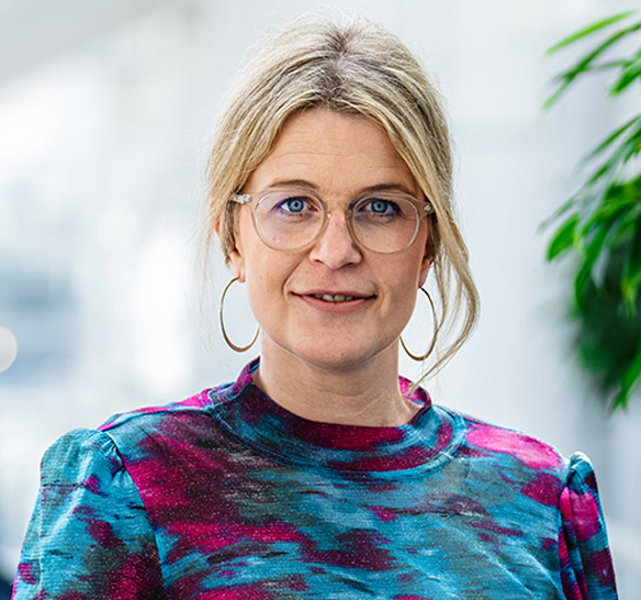 Porträtt av en kvinna med blont hår uppsatt i en lös knut, stora runda glasögon och stora ringörhängen. Hon bär en färgstark tröja i blått, rosa och lila. I bakgrunden syns ett ljust, modernt inomhusutrymme med gröna växter.