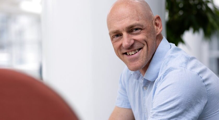 Leende kortklippt man i ljusblå skjorta sitter i ljus, modern inomhusmiljö.