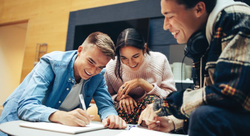 Tre unga personer sitter vid ett lågt bord och samarbetar över anteckningar. Alla ler och verkar engagerade i uppgiften.