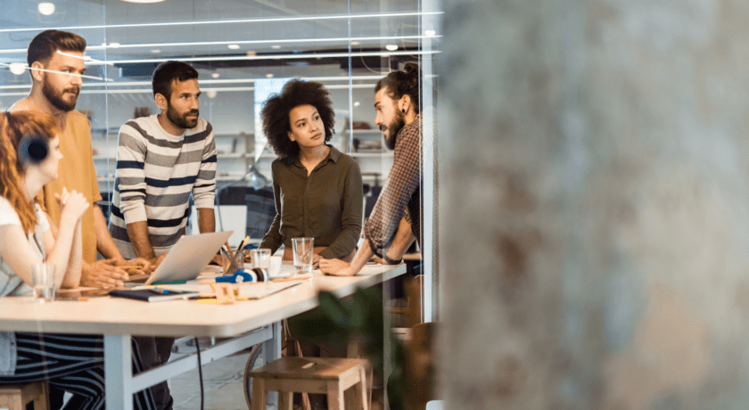En grupp unga vuxna samlade runt ett bord i ett modernt kontorsutrymme. De diskuterar och samarbetar i en kreativ arbetsmiljö, med pennor, glas och en laptop på bordet.