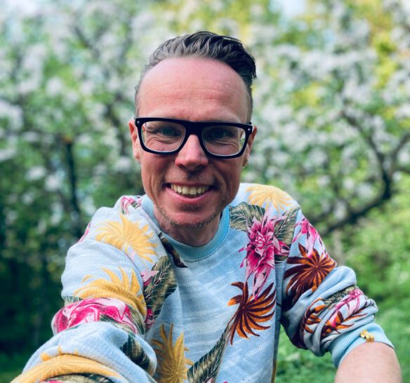 Man i svarta glasögon med ett stort leende, färgglad tröja med blommor på. Bilden är tagen utomhus och har grönska i bakgrunden