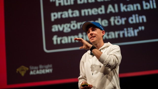 En föreläsare står på scen och gestikulerar engagerat medan han presenterar om AI inför publik. Bakom honom syns en stor presentationsskärm med text om AI och framtida karriärmöjligheter.