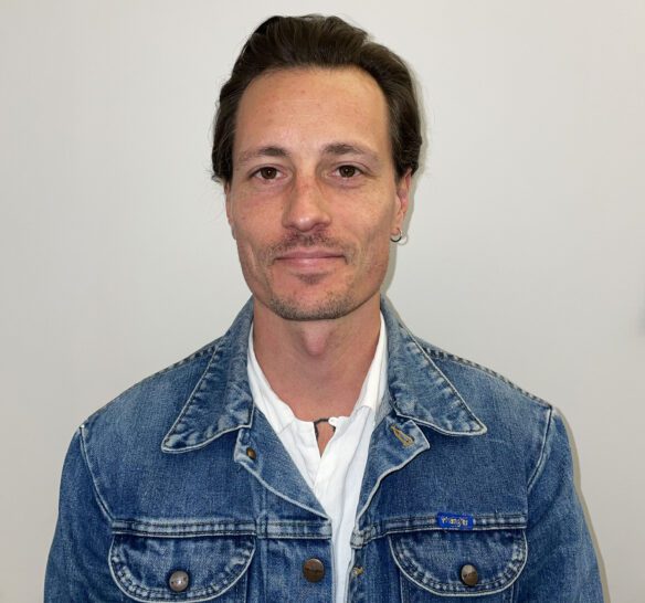 Man som ler lite lätt med mörkt kort hår. Han har en jeansjacka med en vit tröja under på sig och en ljus bakgrund.