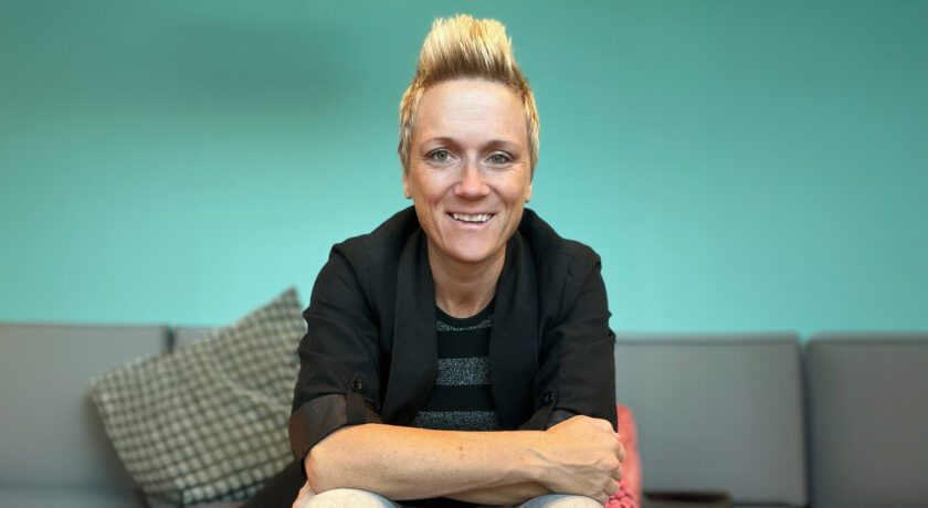 Porträtt av Marlene Kanto som sitter framåtlutad och ler mot kameran, med kort blont hår och svart kavaj mot en mjukt turkos bakgrund.