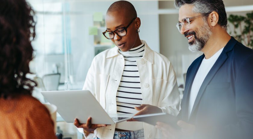 Tre personer står i ett ljust kontor och tittar tillsammans på en laptop. En person håller i datorn och visar något, medan de andra lyssnar och följer med i samtalet. Glasväggar och post-it-lappar syns i bakgrunden.