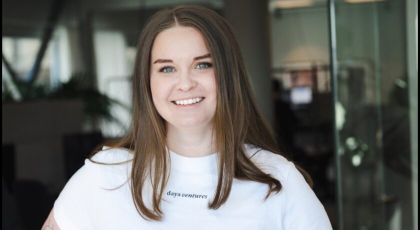 Porträtt av en leende kvinna med långt brunt hår, klädd i en svart tröja med texten “daya ventures”, hon står i ett kontor som syns i bakgrund.