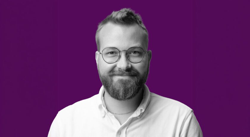 Porträtt i svartvitt av en person med kort hår, skägg och runda glasögon, klädd i ljus skjorta. Personen ler lätt mot kameran. Lila bakgrund.