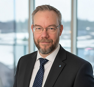 Timo Lagerbjelkes porträtt, en man i glasögon, mörk kavaj, vit skjorta och blå slips ler in i kameran och i bakgrunden är det stora fönsterpartier.