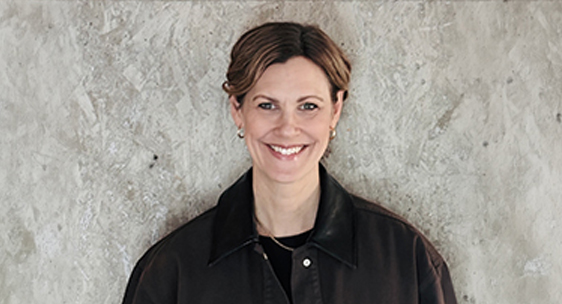 Porträtt av en leende kvinna med uppsatt hår, klädd i en mörk jacka, stående mot en ljus betongvägg.