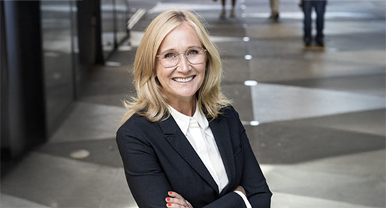 Kvinna med blont mellanlångt hår och glasögon står med armarna i kors och klädd i svart kavaj och vit skjorta och ler in i kameran.