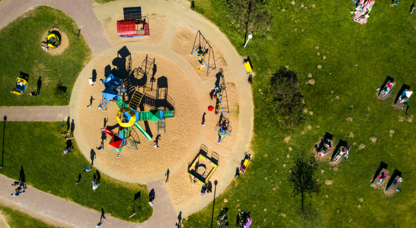 Flygbild över en lekplats i en grön park. I mitten finns färgglada lekredskap omgivna av sand och gångvägar. Barn och vuxna rör sig runt lekplatsen och flera personer sitter eller ligger på gräset i solskenet.
