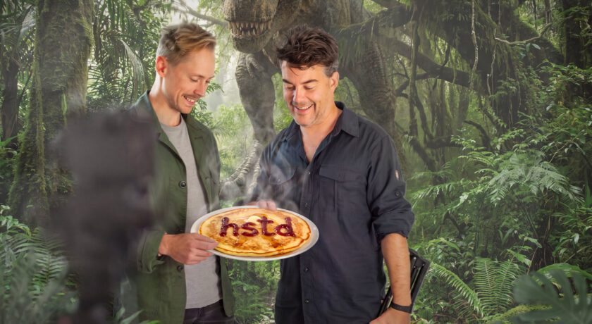 Två killar står i en skog med en dinosaurie bakom och håller i en tallrik med en pannkaka på, med texten hstd skrivet i röd sylt, bilden är AI genererad