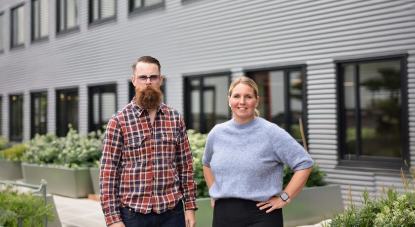 Två personer som står framför en stor byggnad med en del växter. En kille i rutig skjorta, glösögon och skägg bredvid en kvinna med uppsatt hår och blå tröja.