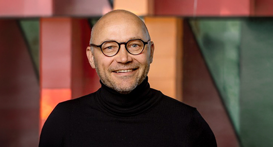 Porträtt av Martin Högenberg som ler mot kameran, med runda glasögon och svart polotröja mot en bakgrund i varma färger.